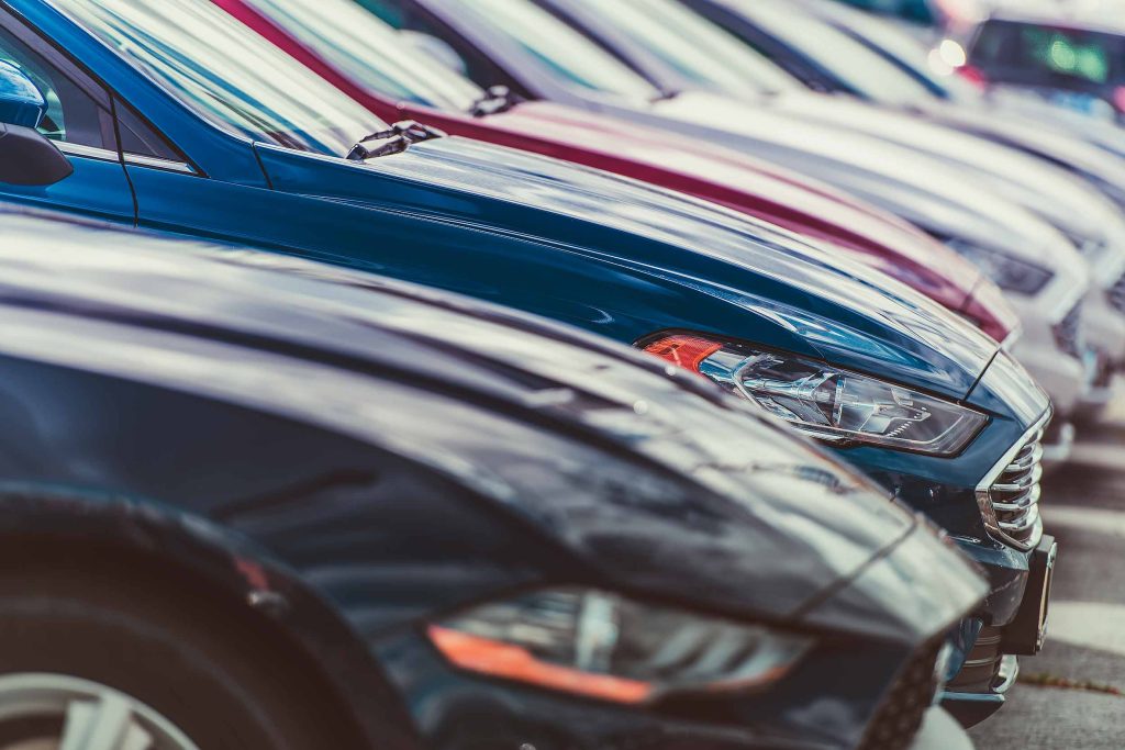 Front ends of several parked cars lined up in a parking lot, with a blue car in the foreground.