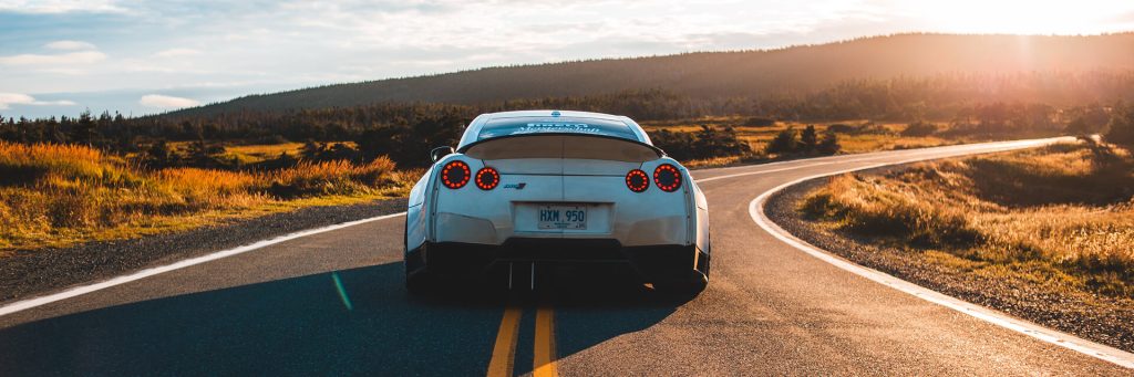 A silver sports car drives on a winding rural road, surrounded by grass and hills under a bright, sunlit sky.