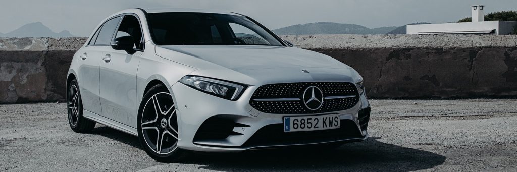 A silver Mercedes-Benz car parked near a weathered concrete wall, with mountainous landscape in the background and cloudy skies above.