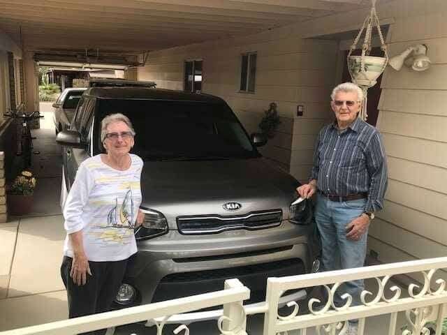 Two persons stand beside a parked Kia car in a sheltered garage. The area appears residential with decorative railings and hanging plants.