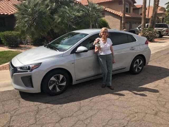 A person stands beside a silver car holding a small dog, in a suburban neighborhood with palm trees and stucco-roofed houses.
