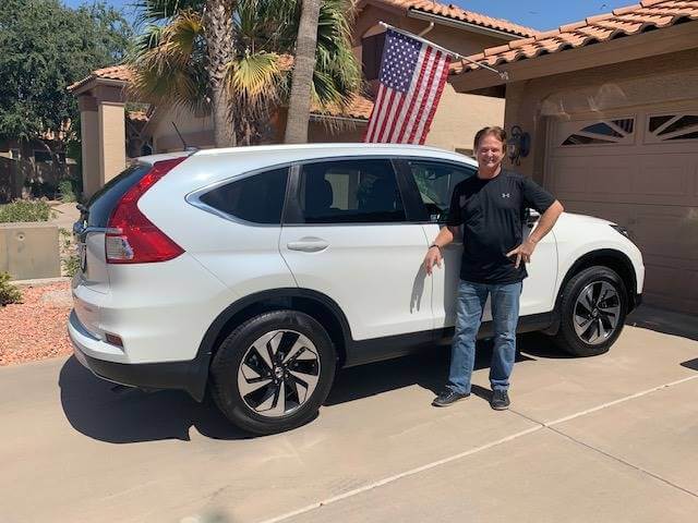 A person stands beside a white SUV on a sunny driveway. An American flag hangs from the house in the background.