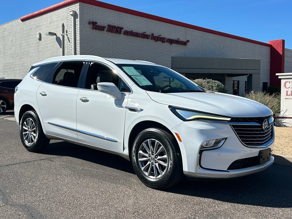 7 White crossover SUV parked in front of a car dealership on a sunny day.